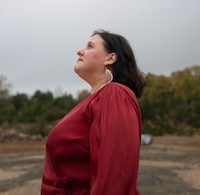 Woman in red dress standing in a rock quarry with trees in the background staring up at the sky